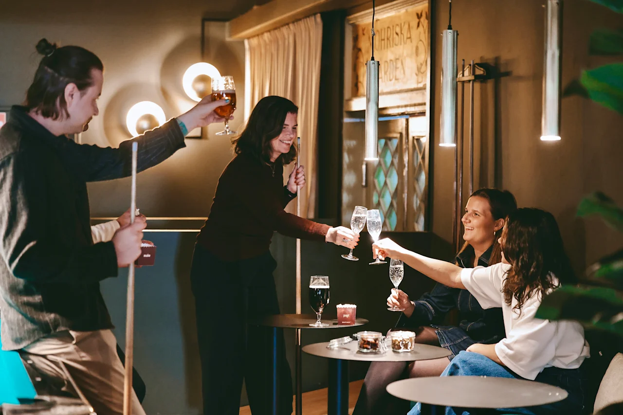 Fem personer skålar med drinkar vid små runda bord, vilket skapar en livlig atmosfär i en varmt upplyst bar. Vägglampor och en biljardkö bidrar till stämningen.