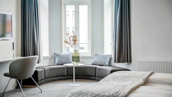 Den välvda grå soffan står nära stora fönster med blå gardiner, vetter mot ett litet vitt bord med en vas med blommor; säng i förgrunden med texturerad filt och TV på väggen.