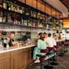 Bartender preparing drinks behind a long, wooden bar. Patrons seated on high stools engage with each other. Shelves stocked with bottles and glasses hang above in a bright, modern bar setting.