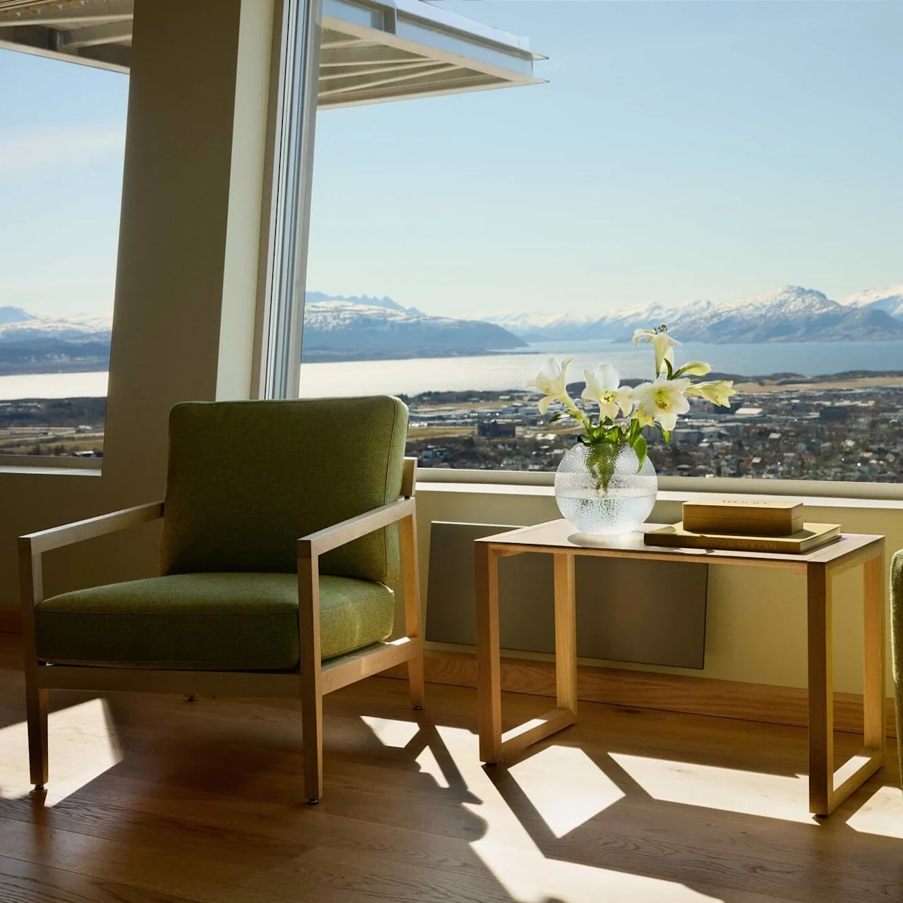 Green armchair sits by a table with a vase of lilies and two books, overlooking a scenic view of mountains and water through large windows.