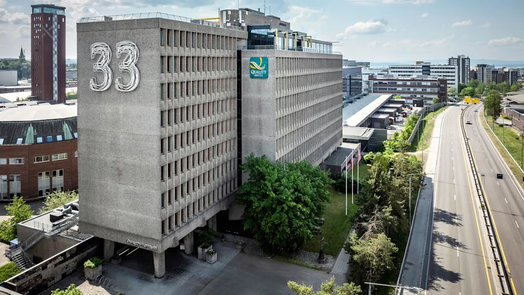 Luftfoto af en stor rektangulær bygning mærket "33" og "Quality Hotel" med tilstødende vej, træer og omkringliggende bylandskab under en delvis overskyet himmel.
