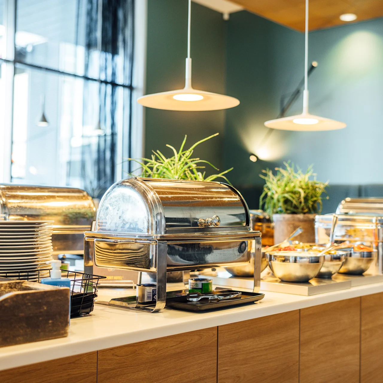 Buffet counter displaying chafing dishes filled with food, surrounded by stacks of plates and potted plants, situated in an airy, modern dining area with pendant lighting.