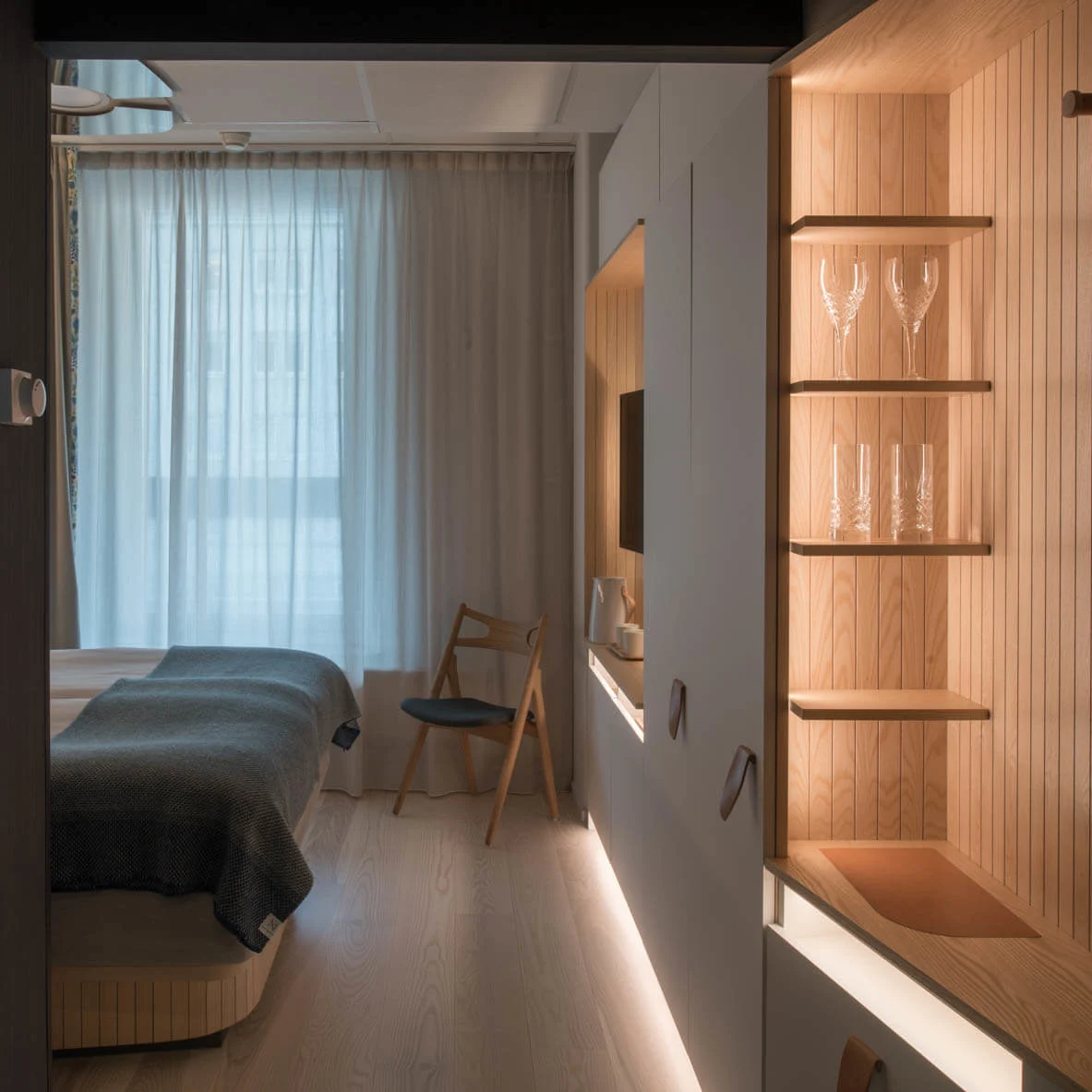 A cozy hotel room features a neatly made bed with a gray blanket, adjacent chair, and wooden shelves holding glasses. Soft lighting and sheer curtains create a serene atmosphere.