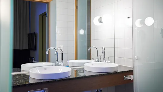 Two white round sinks sit on a dark countertop with chrome faucets. A large mirror and white tiled wall reflect soft lighting; a glass door is partially visible.