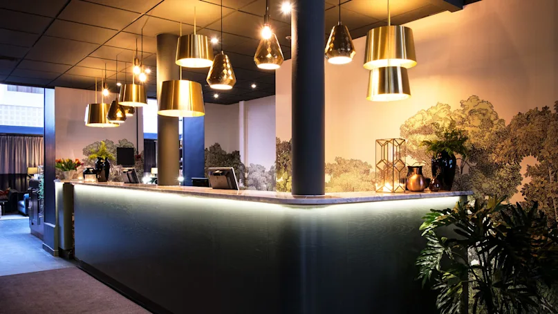 A sleek reception desk with overhead pendant lights in a stylish lobby, featuring decorative plants and a mural.