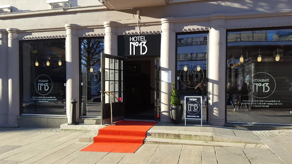 A building with pillars features “HOTEL N°13” signage above a door, flanked by windows showing “RESTAURANT N°13.” A red carpet leads to the entrance, with a sign reading “OPEN! N°13 Hotel. Burgers. WELCOME IN!”