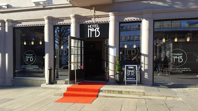 A building with pillars features “HOTEL N°13” signage above a door, flanked by windows showing “RESTAURANT N°13.” A red carpet leads to the entrance, with a sign reading “OPEN! N°13 Hotel. Burgers. WELCOME IN!”