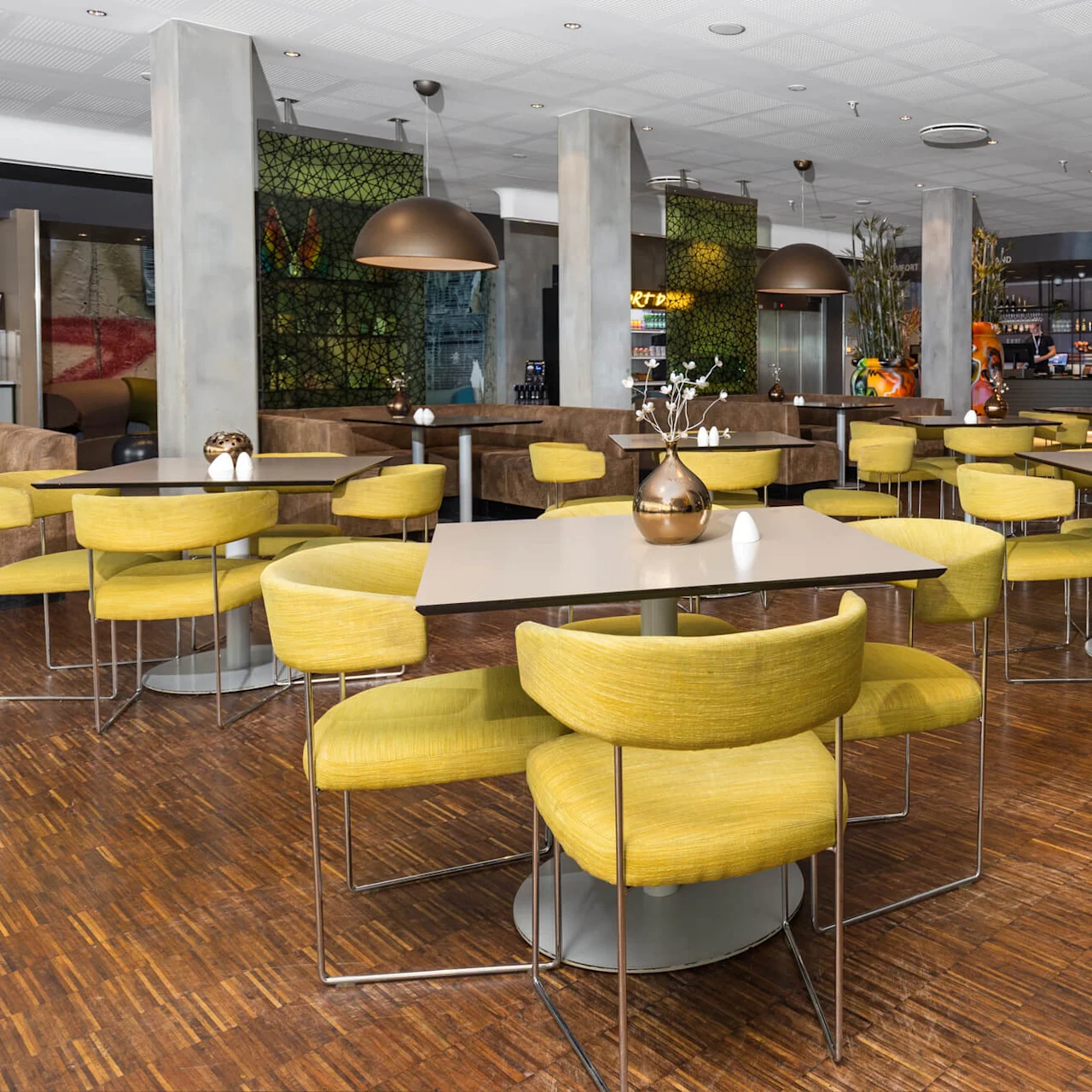 Modern dining area, features yellow cushioned chairs around square tables, under large pendant lights. Wooden floor and decorative plants enhance the cozy atmosphere.