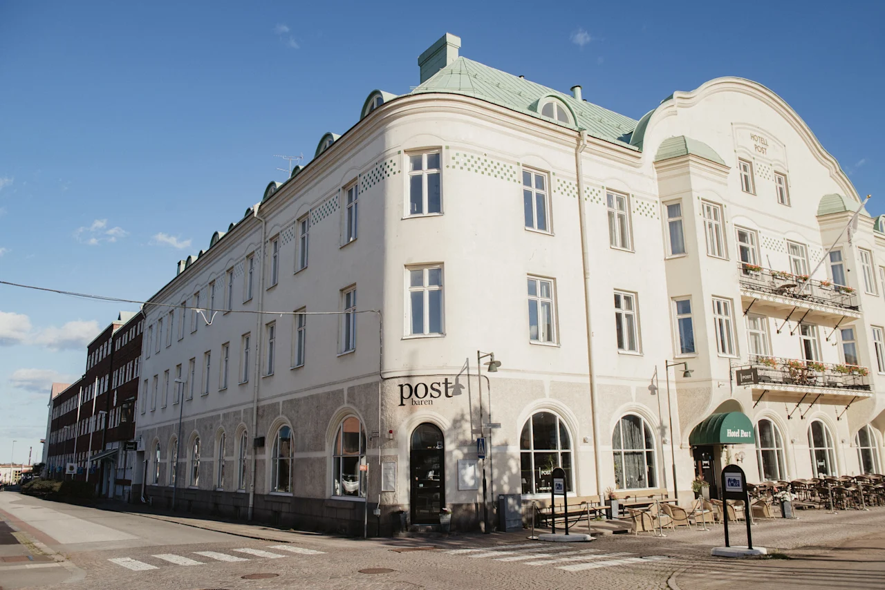 En stor, elegant hotellbyggnad med uteservering en solig dag. Text på byggnaden lyder Hotel Post och post baren.