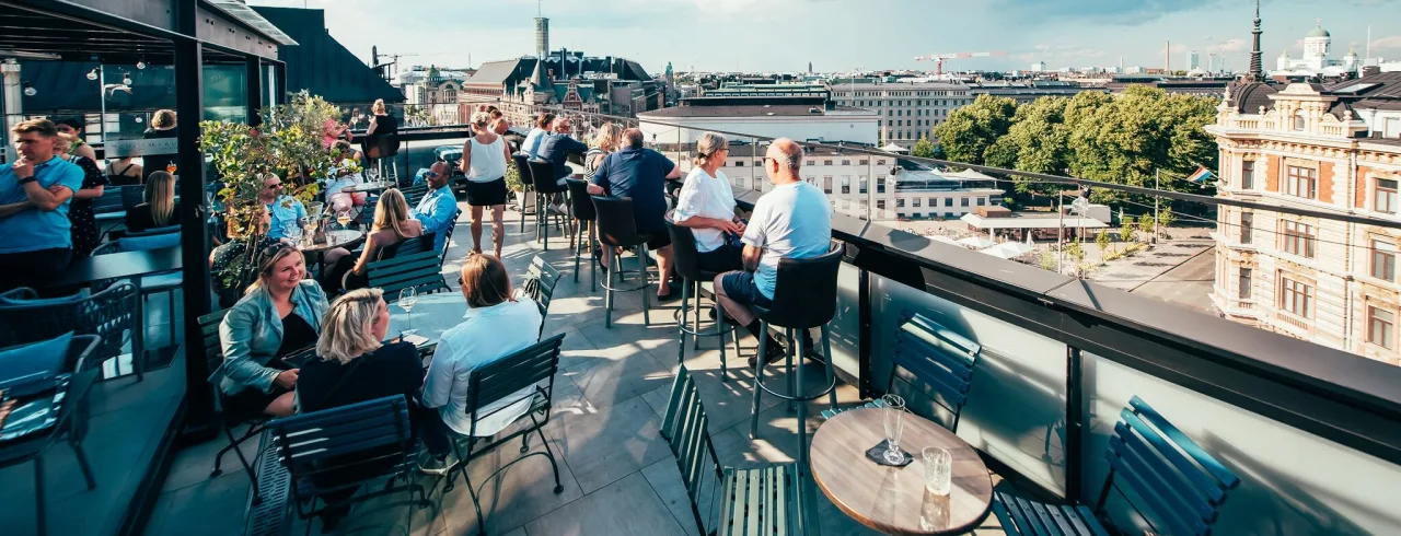 Mennesker sosialiserer på en takbar med utsikt over byens silhuett under en klar himmel.