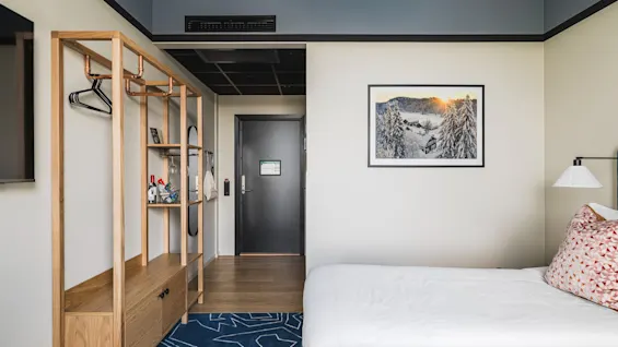 Et moderne hotelværelse med en komfortabel seng, en åben opbevaringsenhed og et indrammet vinterlandskabskunstværk.