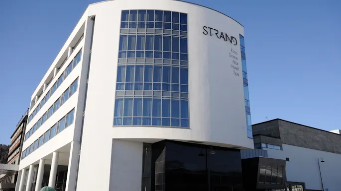 A modern multi-story building with wide glass windows curves upwards under a clear blue sky. "STRAND" is prominently displayed on the facade with smaller text: "Sola Hotel Spa."