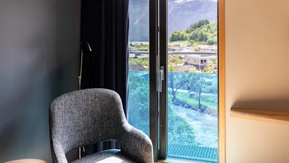 En grå stol står vänd mot ett öppet fönster, med utsikt över träd, berg och blå himmel. Solljus strömmar in och lyser upp interiören.