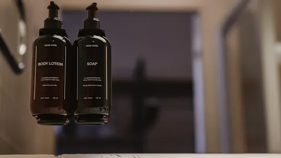 Two bottles labeled "BODY LOTION" and "SOAP" with text “HOME HOTEL”, and descriptions. 280 ml each, 100% vegan, sit on a bathroom shelf against a blurred background.