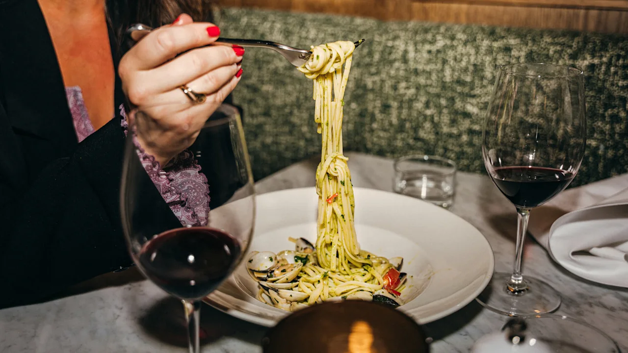 En person snurrer spaghetti med muslinger på en gaffel, ledsaget af to glas rødvin, ved et restaurantbord med en grøn polstret bænk.
