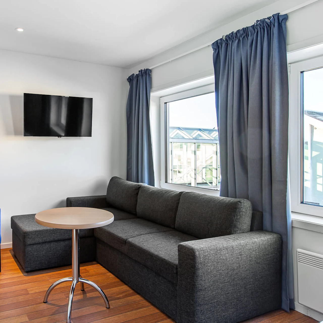 Et moderne oppholdsrom har en mørkegrå sofa med et rundt bord, en blå lenestol, en veggmontert TV og et trebord med lampe, innrammet av store vinduer og gardiner.