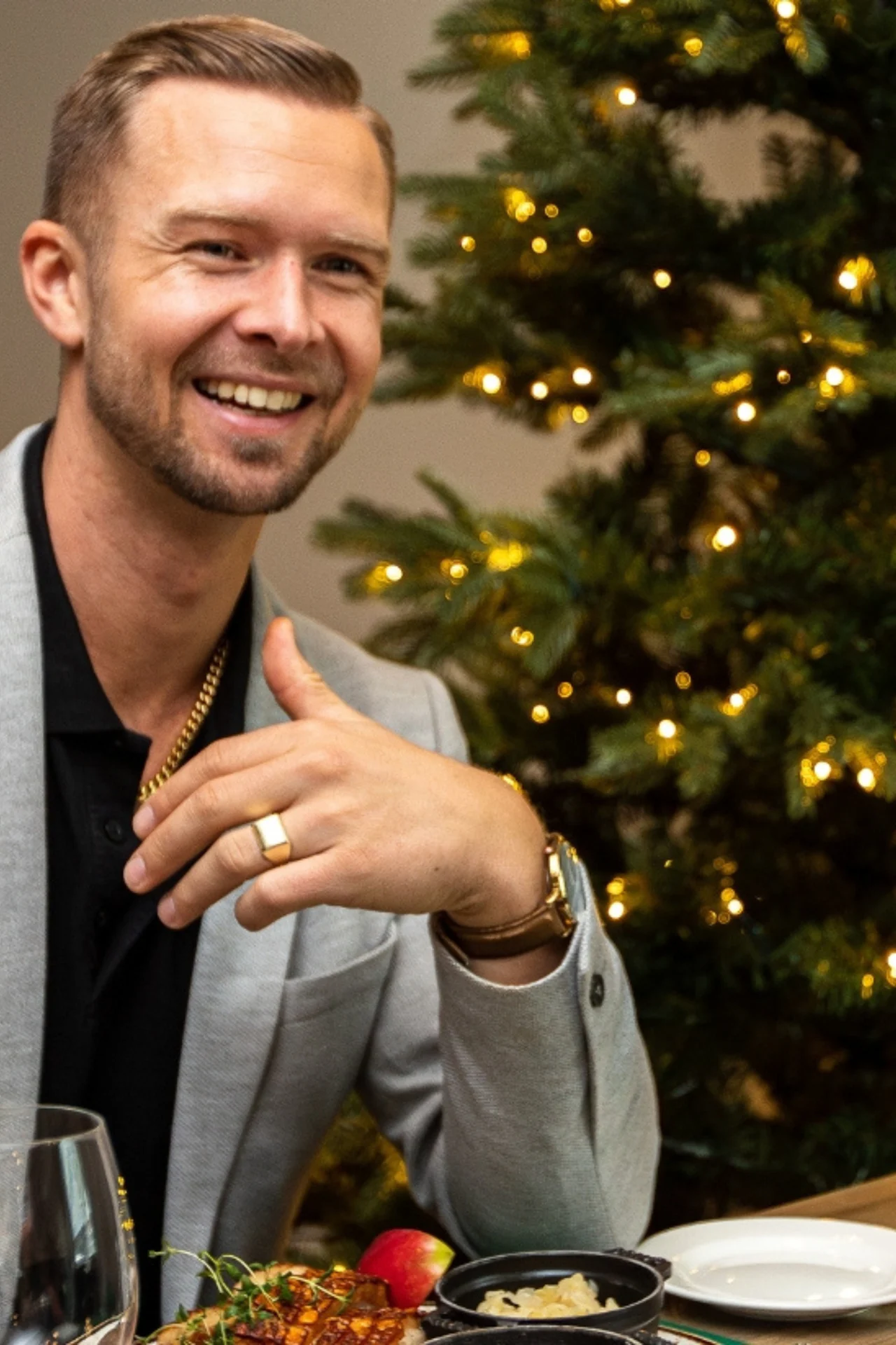 En mann i lys jakke smiler og snakker ved et spisebord, dekorert med festlige tallerkener, nær et opplyst juletre. En annen person er delvis synlig i forgrunnen.