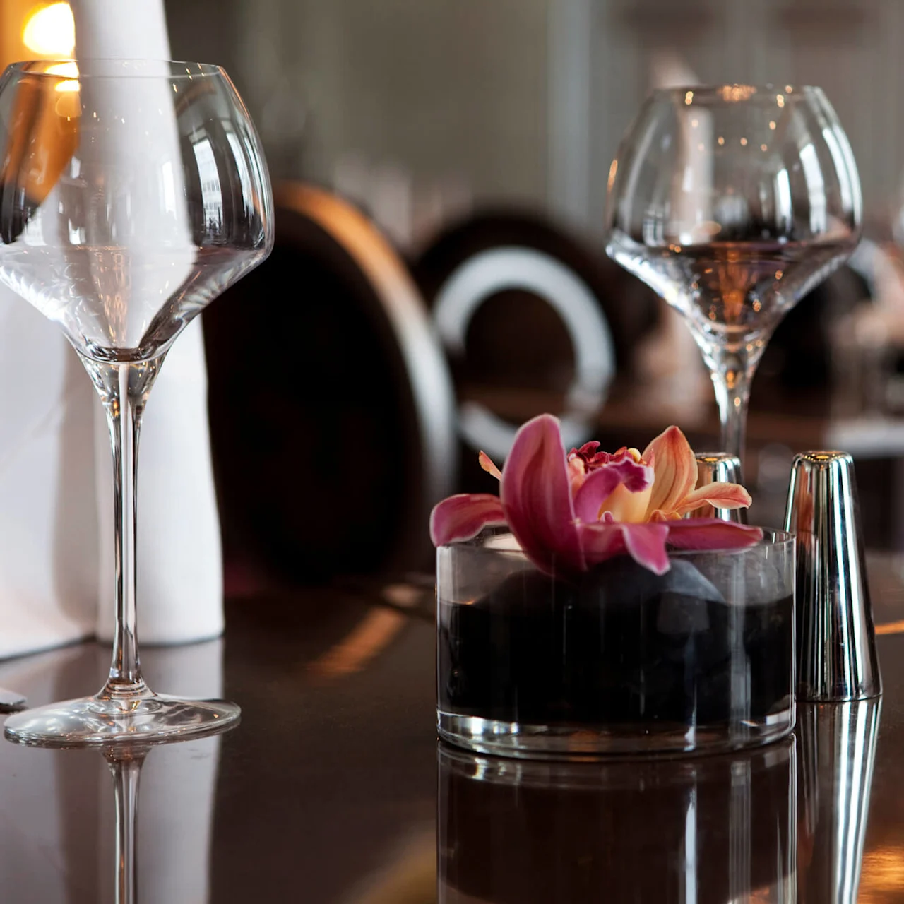 Et vinglas står på et restaurantbord og reflekterer blødt lys. Ved siden af flyder en orkidé i en lille glaskrukke. Andre borde og stole fylder baggrunden.