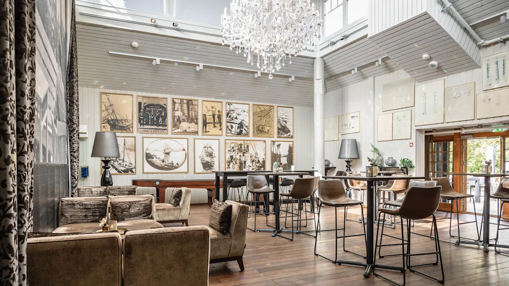 En lyxig ljuskrona hänger ovanför en elegant lounge med vintagefotografier på väggarna, bruna stoppade stolar runt borden. Ljust naturligt ljus filtreras genom stora fönster och skapar en sofistikerad, luftig atmosfär.