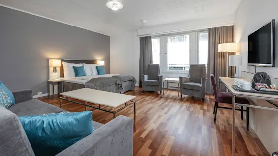 Seng med turkis puder mod en grå væg, ved siden af et godt oplyst opholdsområde med moderne møbler, flere stole, borde og store vinduer, under et hvidt loft i et rummeligt rum.