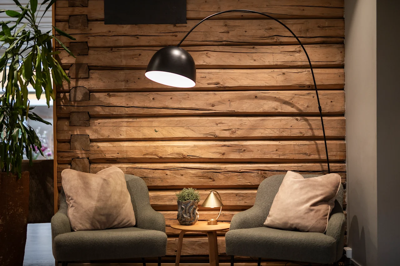 To lænestole med puder vender mod hinanden, adskilt af et lille bord med en plante og lampe. En buet gulvlampe lyser op mod en trævæg.
