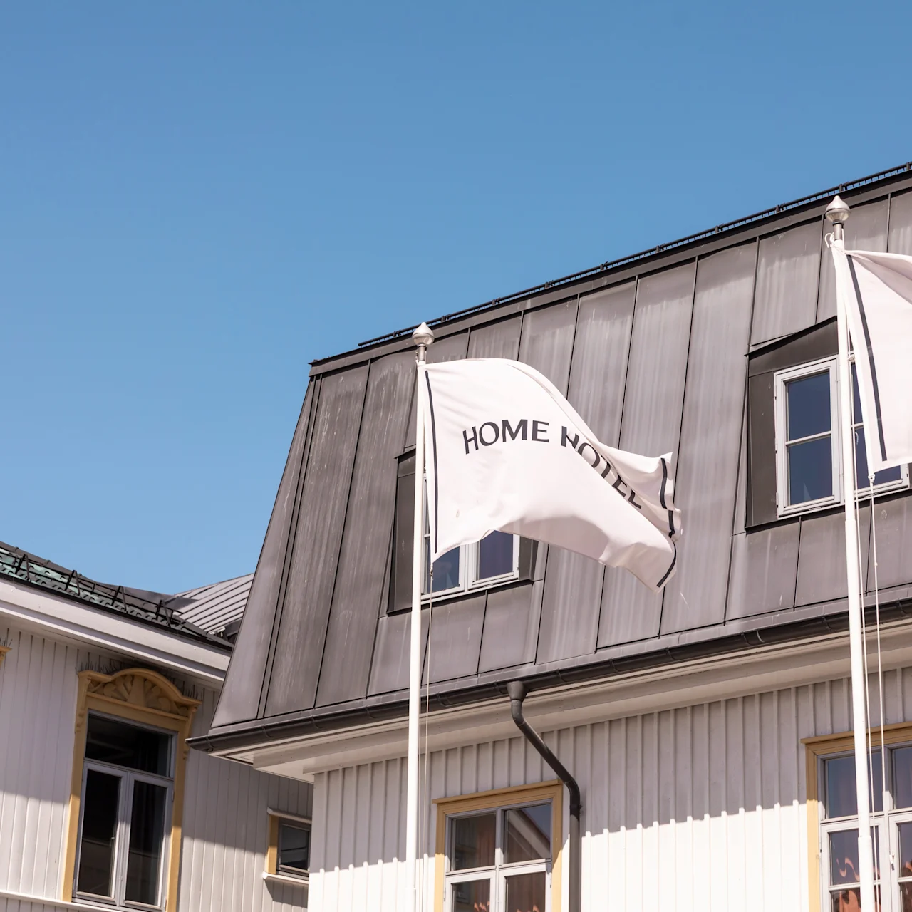 To flag markeret "HOME HOTEL" blafrer på stænger foran en bygning med skråt tag og flere vinduer under en klar blå himmel.