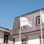 To flag markeret "HOME HOTEL" blafrer på stænger foran en bygning med skråt tag og flere vinduer under en klar blå himmel.