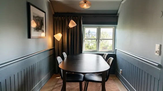 Et mørkt træbord med fire stole står under en loftlampe. Ved siden af oplyser en trehovedet gulvlampe et indrammet billede på væggen i et smalt, blødt oplyst rum.