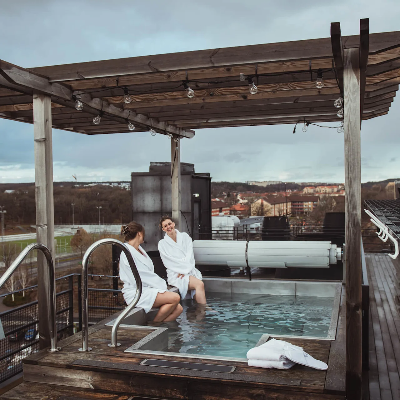 Two people wearing white robes relax in a rooftop hot tub under a wooden pergola, overlooking an urban landscape and sports field.