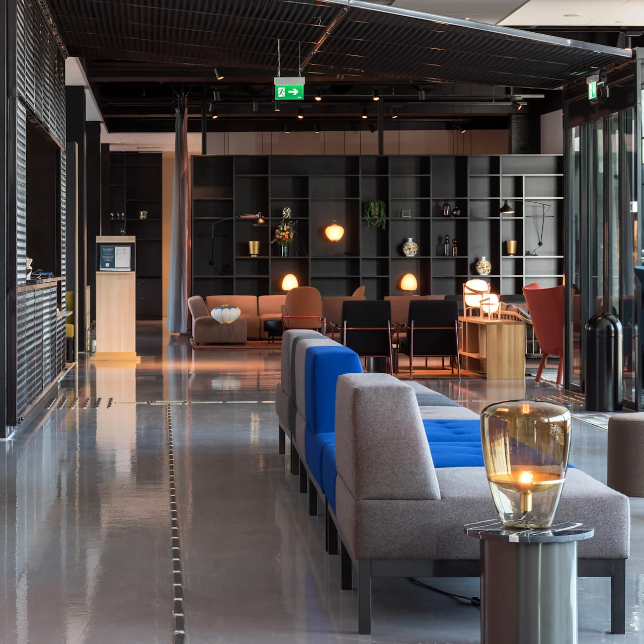 A modern lounge features colorful seating, a side table with a lamp, and a coffee machine on the wall. Shelves with plants and lights in the background. Exit signs visible.