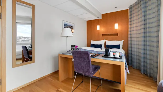 Et pent organisert hotellrom med en dobbeltseng med puter, et skrivebord med stol og lampe i et rom med tregulv. Et speil reflekterer en vindusutsikt mot en kystlinje.