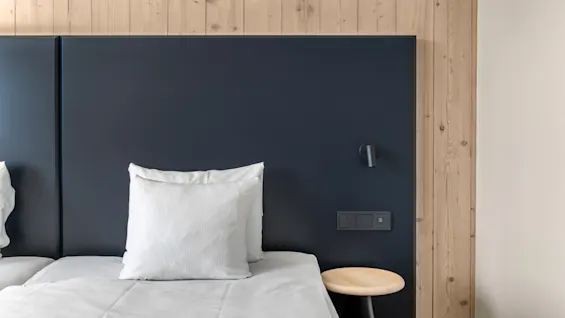 Et moderne hotelværelse med en seng, mørk sengegavl, træpaneler på væggen og et lille sidebord med lampe og kontakter.