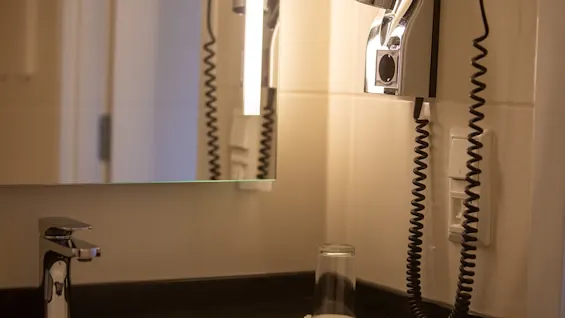 En veggmontert hårføner henger ved siden av et speil og reflekterer lys på et bad. Nedenfor hviler en kromkran og et opp-ned glass på en mørk benkeplate.