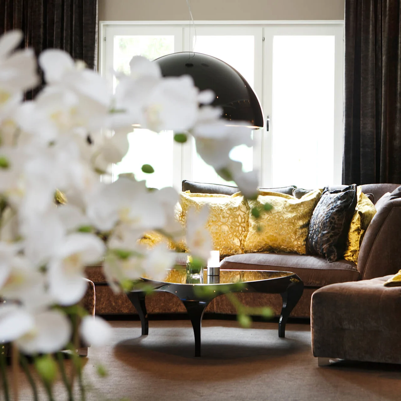 An elegantly decorated living room with plush sofas, patterned cushions, and large windows, accentuated by soft lighting and ornate flowers.