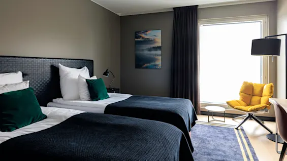 Two neatly made beds with green pillows face a large window. A cozy yellow chair and small table sit beside, with a painting of a misty lake on the wall.