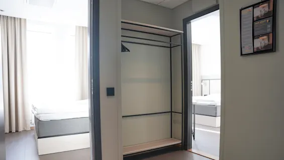 Et rom med en tom metallramgarderobe står i et hjørne. Sollys lyser opp to tilstøtende rom med senger og beige gardiner.