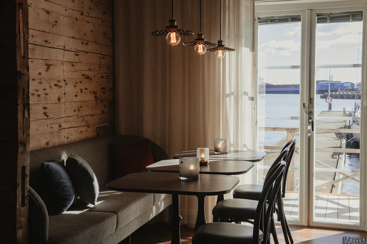 Et hyggeligt indendørs siddeområde med trævæg, sofa, borde og stole, med udsigt over et roligt vandområde og en anløbsbro.