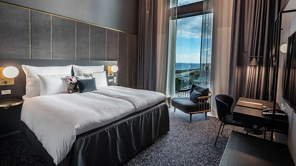 A large bed with white linens is positioned beside a floor-to-ceiling window, revealing an ocean view. The room includes a desk, chair, and modern lighting.
