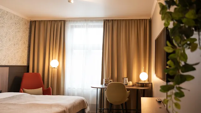 Et pent organisert hotellrom har en seng og en skrivebordstol. Varm belysning lyser opp et delvis dekket vindu, med en TV og en grønn plante på veggen.