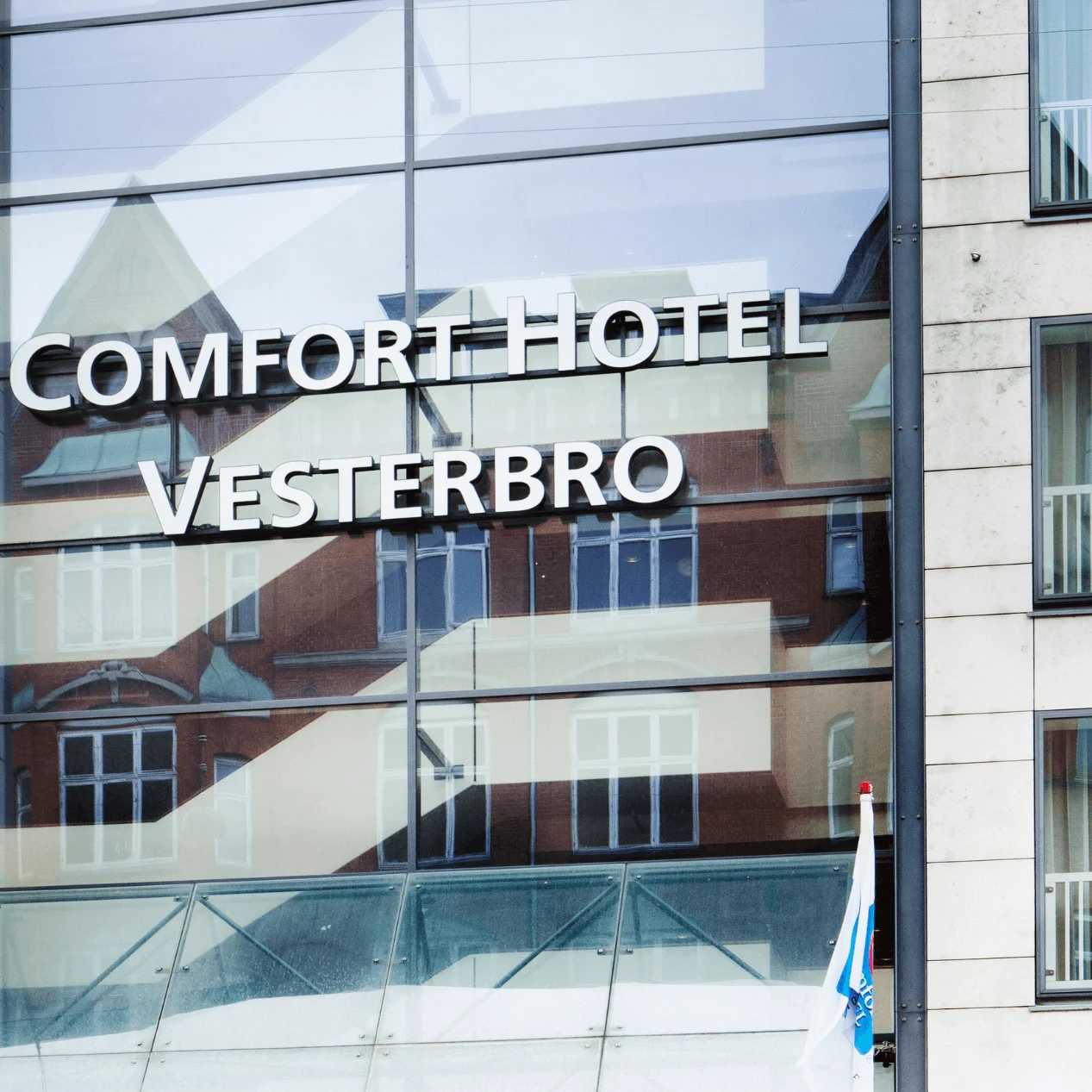Comfort Hotel Vesterbro sign displayed on a modern glass building facade, reflecting nearby architecture. Two small flags are visible below. 