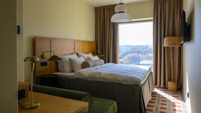 A cozy hotel room with a neatly made bed in the center, flanked by two lamps. Large window with cityscape view, surrounded by beige curtains.