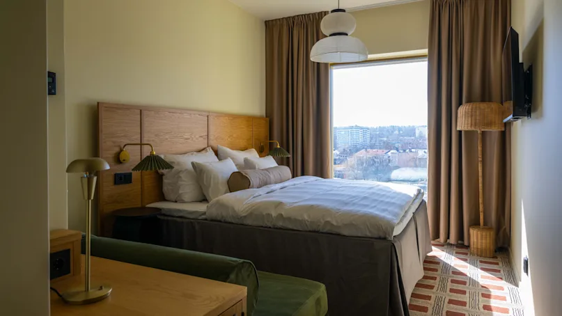 A cozy hotel room with a neatly made bed in the center, flanked by two lamps. Large window with cityscape view, surrounded by beige curtains.