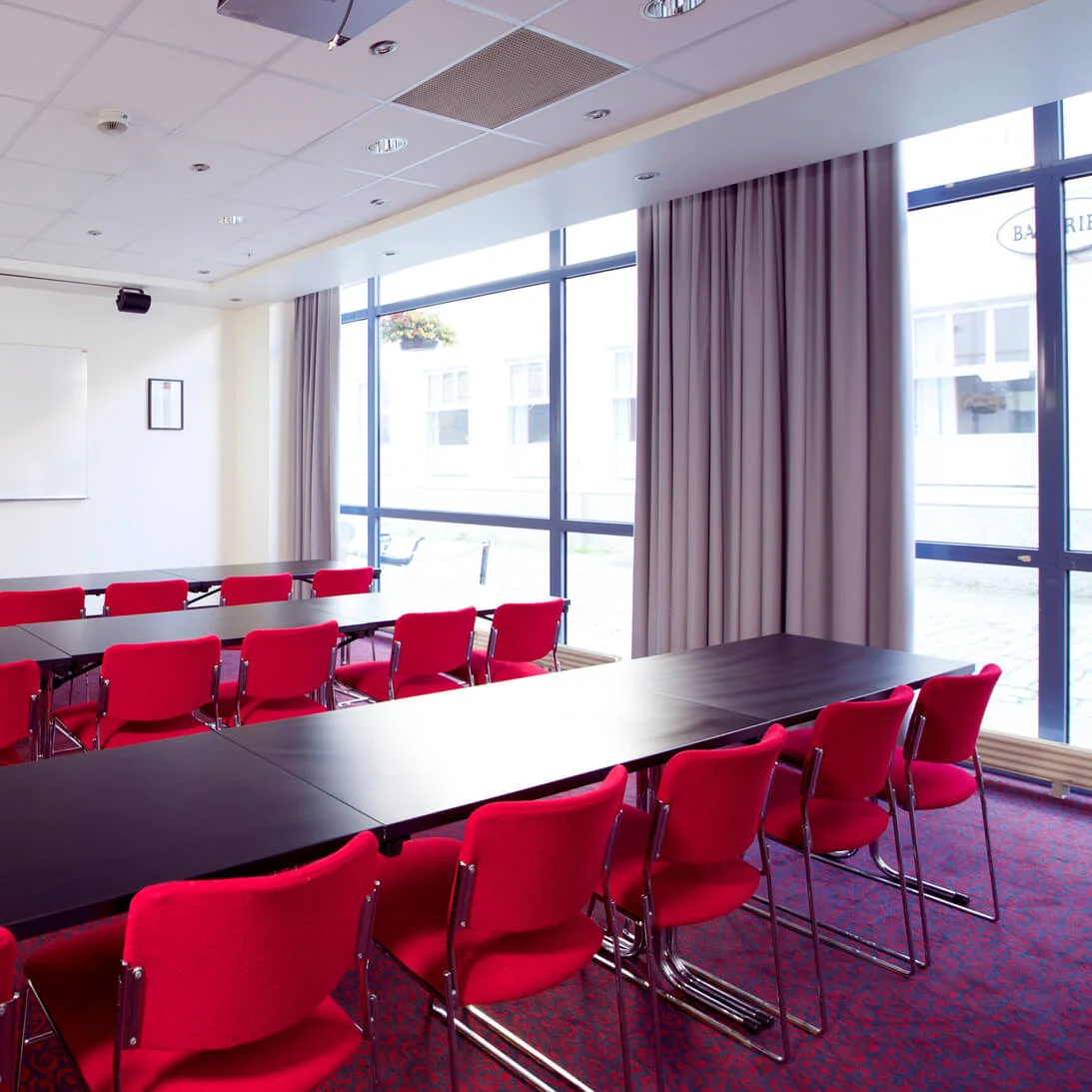 A conference room with red chairs, a long table, whiteboard, and projector, brightly lit with natural light from large windows.
