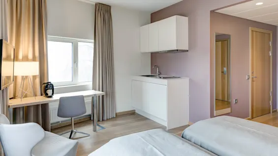 A compact hotel room with a bed, kitchenette, and desk beneath a window, featuring light curtains and a lamp on the desk.