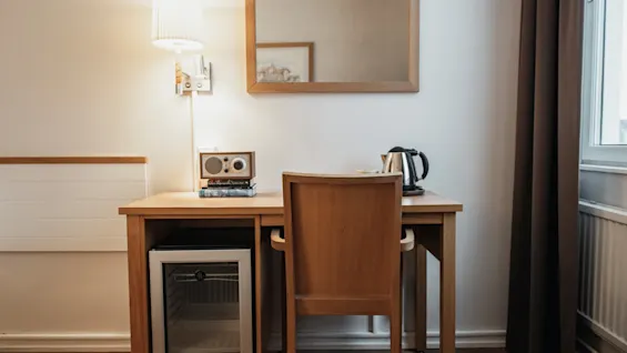 Ett inbjudande hotellrumshörn med skrivbord, stol, lampa, radio, vattenkokare och minikylskåp.