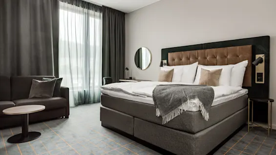 En pænt redt seng med puder og et gråt tæppe er i et hotelværelse, ledsaget af en sofa, sideborde og et stort vindue med gardiner.