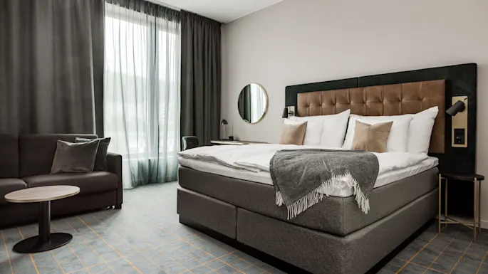 En pent oppredd seng med puter og et grått teppe er i et hotellrom, ledsaget av en sofa, sidebord og et stort vindu med gardiner.