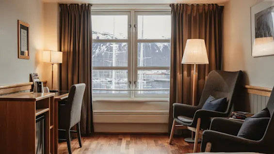 A cozy hotel room with a stunning view of a snowy mountain and water. Features a comfortable workspace and seating area.