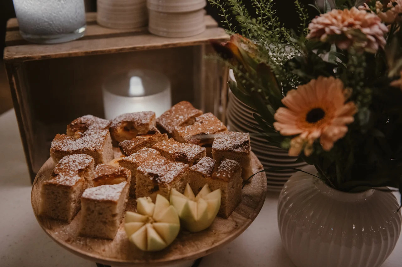 En træplade med flormelisdækkede kagestykker og udskåret æble, ved siden af en vase med blomster og et glødende lys.