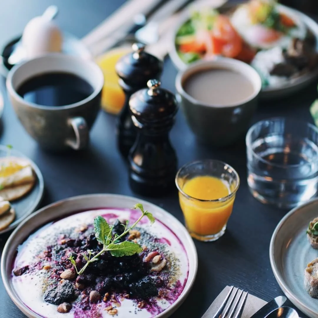 En tallerken med yoghurt toppet med bær og mynte står på et bord omgivet af forskellige morgenmadsretter som croissanter, kaffe, juice og frisk frugt.